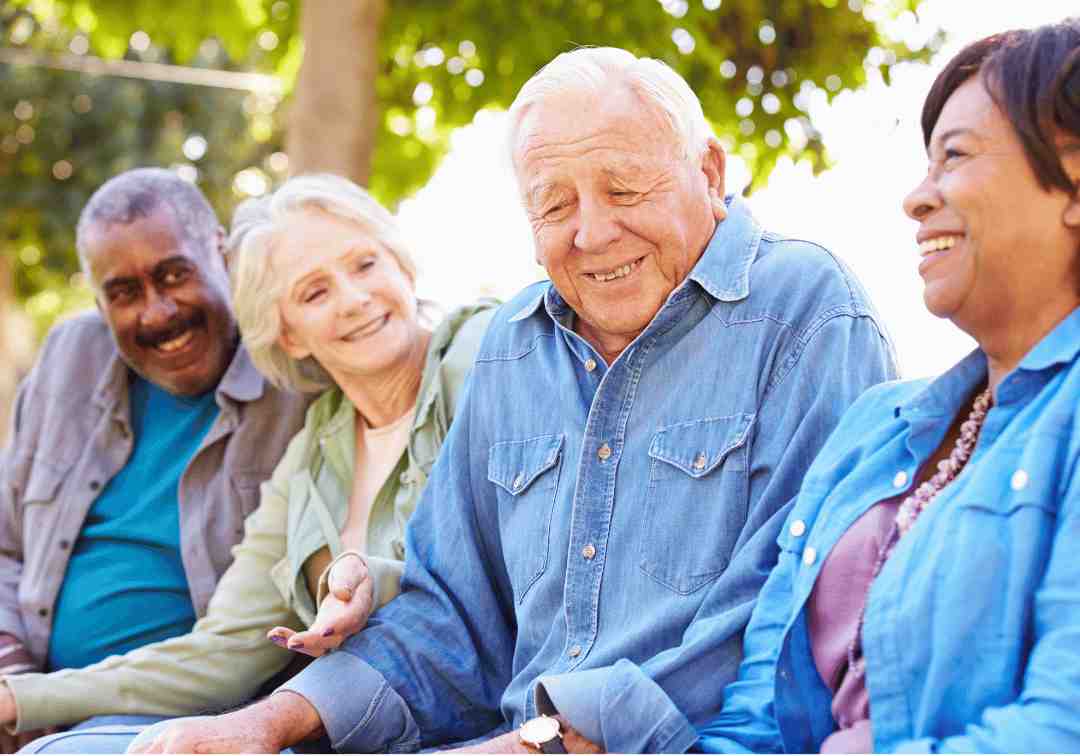 Four senior friends laughing together while sitting outside on a bench.